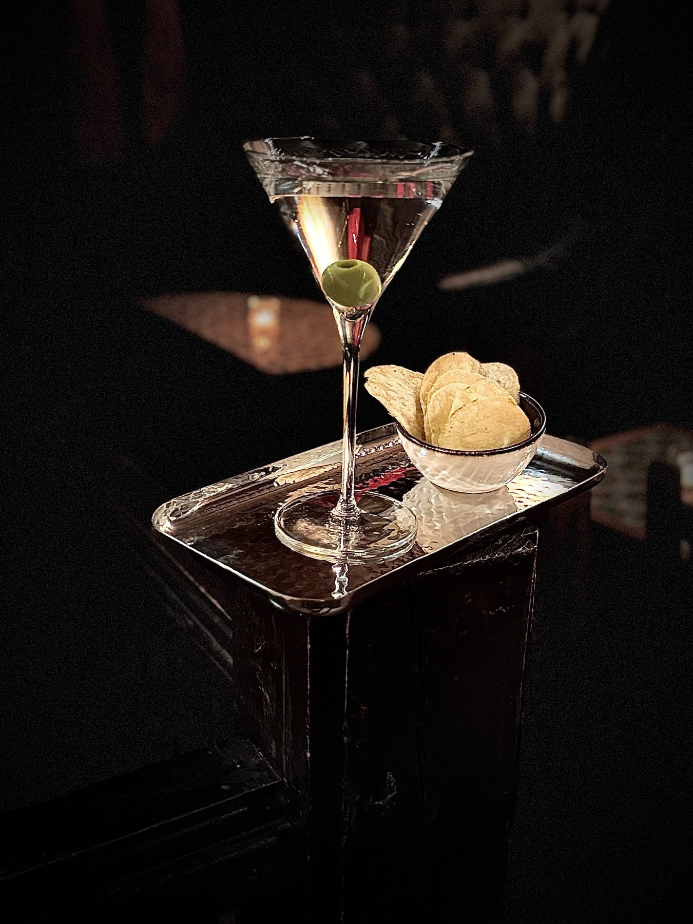 A martini with an olive beside a bowl of chips on a hammered silver tray, lit by candlelight